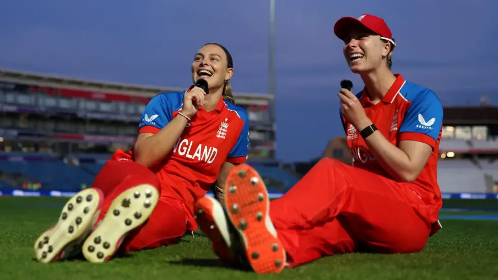 Dani Gibson and Freya Kemp sit together on the outfield, holding mics and smiling
